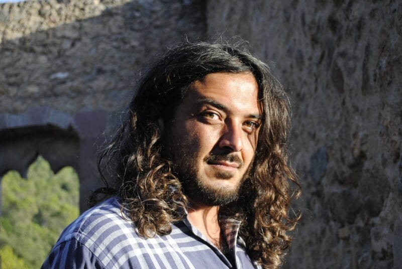 Portrait of a man with long wavy hair in a blue plaid shirt, standing beside a stone wall outdoors, looking at the camera.